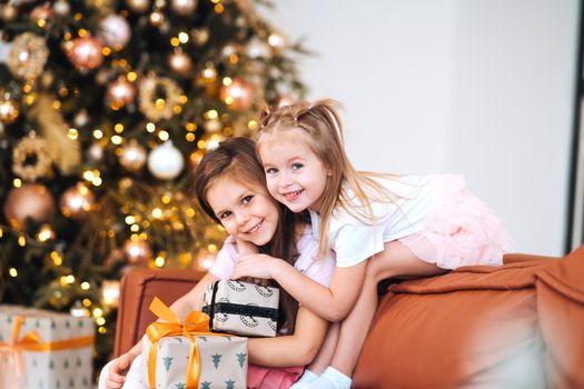 Two little girls are playing with New Year's gifts by the tree at sofa. Christmas, New Year tree.