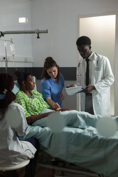 Nurse woman checking oximeter while african american doctor writing medical treatment on clipboard during clinical appointment in hospital ward. Specialist physician monitoring sick patient