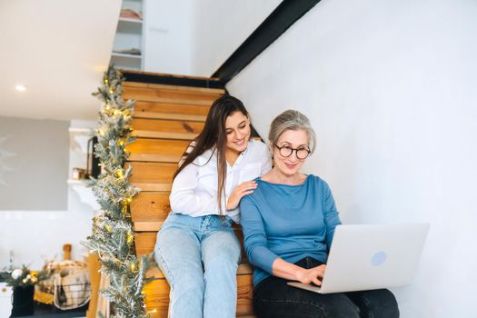 Mother and daughter sitting on the steps and watching something on laptop at home