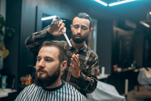 Great time at barbershop. Cheerful young bearded man getting haircut by hairdresser while sitting in chair at barbershop