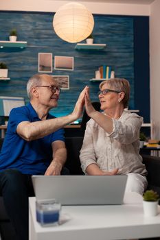 Retired man and woman highfive while using laptop on table at home. Senior married couple enjoying time together with technology while sitting on living room sofa. Old cheerful people