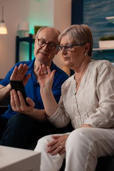 Retired man and woman chatting on video call conference with friends and family while sitting on living room couch. Aged couple using online remote communication together at home