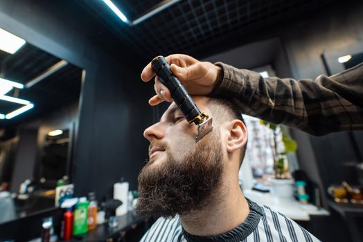 Man visiting hairstylist in barbershop. Barber works with a beard clipper. Hipster client getting haircut.