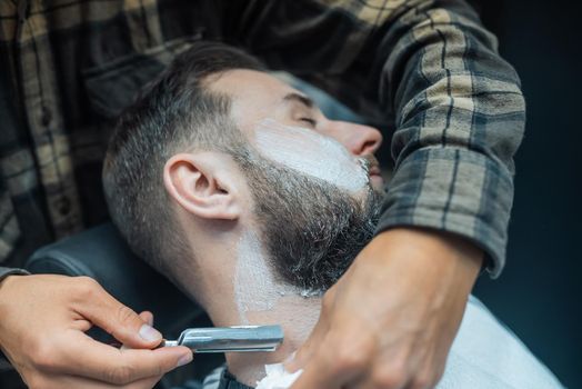 Young bearded man getting shaved with straight edge razor by hairdresser at barbershop