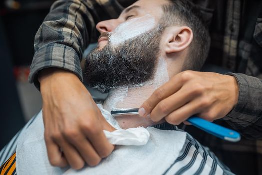 Young bearded man getting shaved with straight edge razor by hairdresser at barbershop