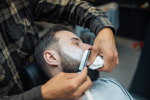 Young bearded man getting shaved with straight edge razor by hairdresser at barbershop