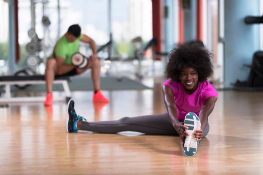 happy young african american woman in a gym stretching and warming up before workout young mab exercising with dumbbells in background