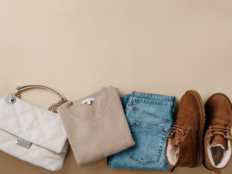 Stylish monochrome winter outfit set. Cashmere knitted beige sweater, blue jeans, chestnut brown chukka suede boots with wool fur and neutral beige bag on champagne neutral background. Top view or flat lay. Copy space