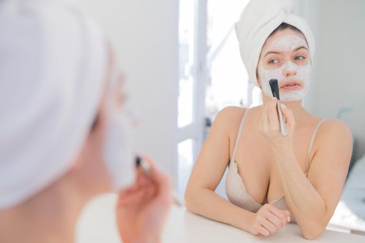 Beautiful caucasian woman with a towel on her hair applies a clay face mask. Taking care of beauty at home.