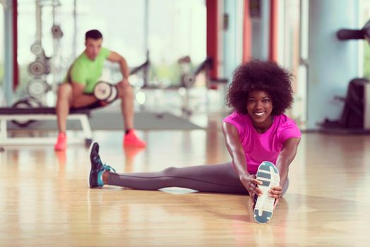 happy young african american woman in a gym stretching and warming up before workout young mab exercising with dumbbells in background