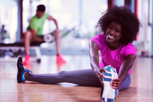 happy young african american woman in a gym stretching and warming up before workout young mab exercising with dumbbells in background