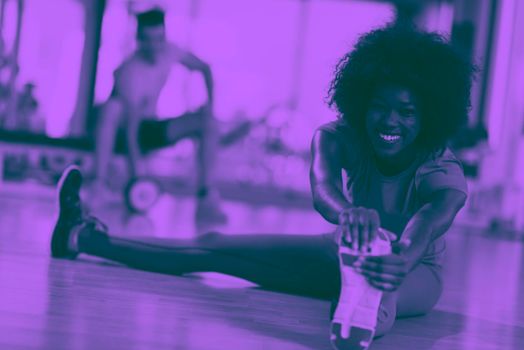 happy young african american woman in a gym stretching and warming up before workout young mab exercising with dumbbells in background duo tone filter