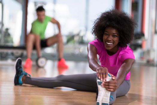 happy young african american woman in a gym stretching and warming up before workout young mab exercising with dumbbells in background