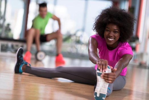 happy young african american woman in a gym stretching and warming up before workout young mab exercising with dumbbells in background