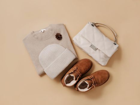 Stylish monochrome winter outfit set. Cashmere knitted bini hat, cashmere beige sweater, chestnut brown chukka suede boots with wool fur and neutral beige bag on champagne neutral background. Top view or flat lay. Copy space
