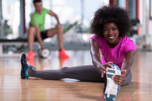 happy young african american woman in a gym stretching and warming up before workout young mab exercising with dumbbells in background