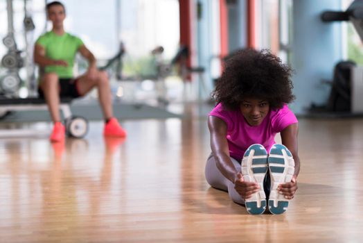 happy young african american woman in a gym stretching and warming up before workout young mab exercising with dumbbells in background