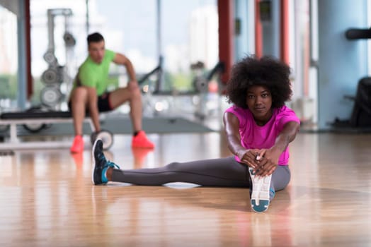 happy young african american woman in a gym stretching and warming up before workout young mab exercising with dumbbells in background