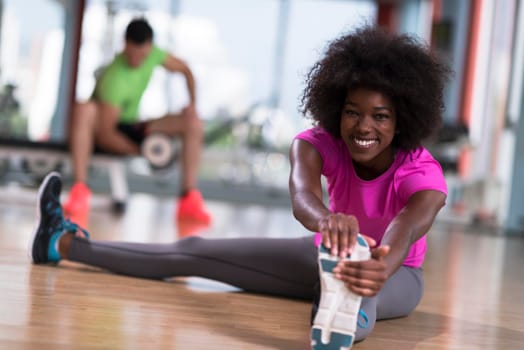 happy young african american woman in a gym stretching and warming up before workout young mab exercising with dumbbells in background