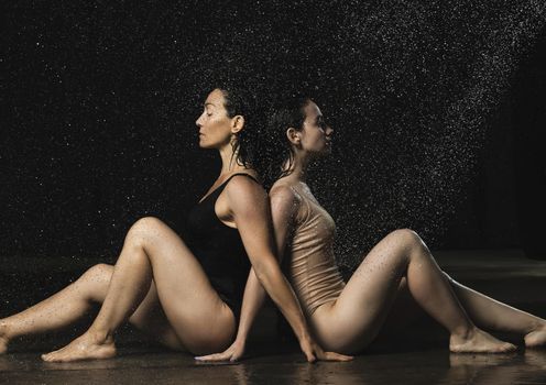 two beautiful women of Caucasian appearance with black hair sit back to back in drops of water on a black background. Women with sad faces