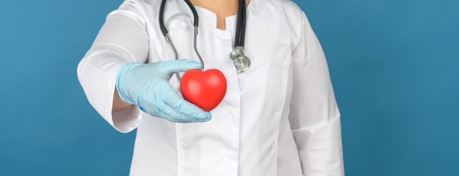 female medic in a white coat stands and holds a red heart on a blue background, the concept of donation and kindness