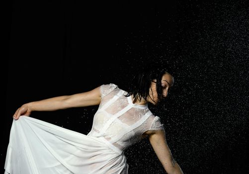 beautiful woman of Caucasian appearance with black hair dances in drops of water on a black background. The woman is wearing a white chiffon dress