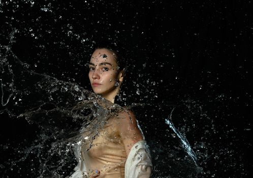 beautiful young woman of Caucasian appearance with black hair stands in drops of water on a black background. Woman wearing white shirt and looking at the camera, serious face