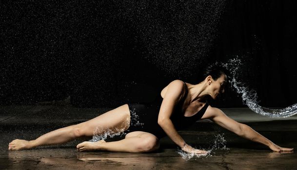 young woman with black hair lies on the ground under raindrops on a black background. Woman dressed in black bodysuit