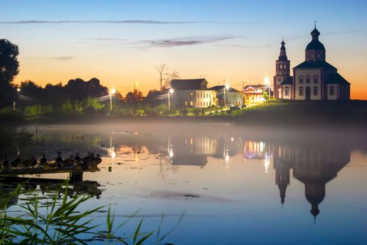 Beautiful evening sunset of a small town reflected in the lake. Night fog over the lake.