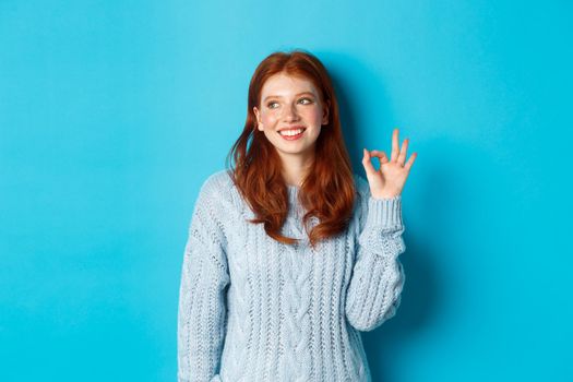 Image of beautiful redhead woman showing okay sign and smiling satisfied, like and agree, standing against blue background.