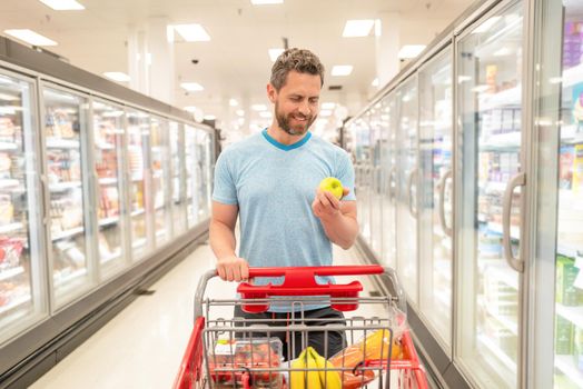 happy man with shopping cart hold apple. customer consumer with purchases. guy at grocery store. shopper at groceries. buying food at supermarket. consumption.