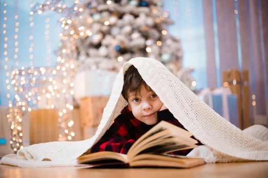 A boy in pajamas reads a book under a Christmas tree . New Year's mood. Reading books . Children's books. A gift for the new year
