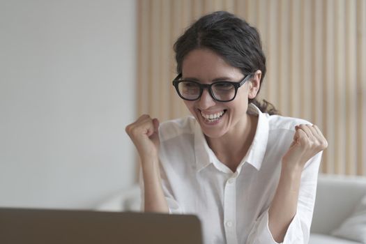 Excited italian woman in glasses looking at laptop screen and celebrating work success, receiving job offer by email, overjoyed female employee clenching fists while getting pleasant news on computer