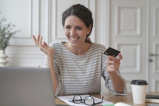 Young smiling italian woman making payment with plastic card during online shopping on laptop at home, sitting at table and looking at computer screen with overjoyed face expression. Ecommerce concept