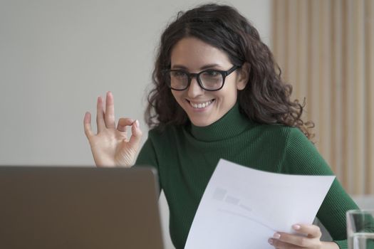 Smiling Italian female employee holding financial report and showing okay gesture during video call with colleagues on laptop computer, happy Spanish business woman in spectacles having web conference