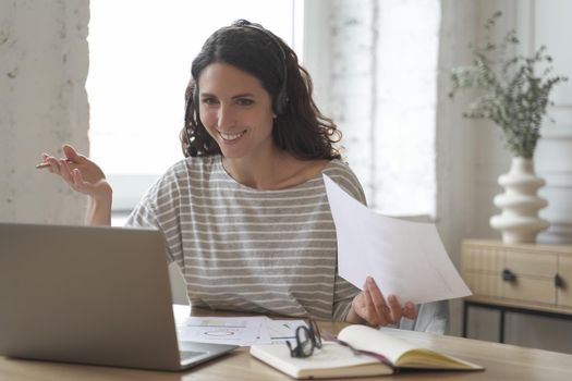 Cheerful smiling female home office employee having video call web conference, looking with excitement at laptop screen, spanish woman wearing headset talking about benefits of new business plan