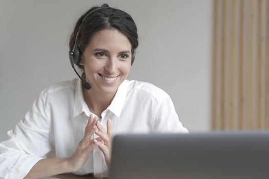 Smiling italian businesswoman in wireless headphones sitting at desk while works on laptop at home office, pleasant confident female employee consults clients online using modern technology