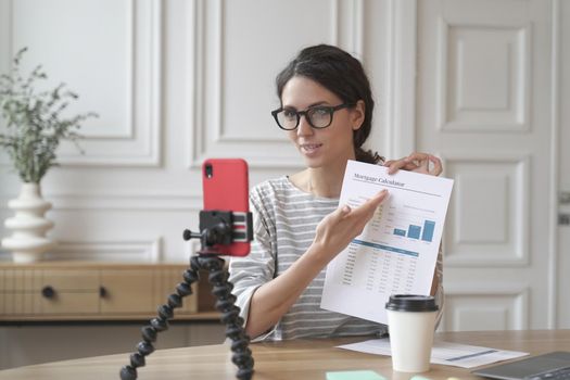 Young hispanic woman real estate advisor talking with client online about monthly mortgage payment, sitting in front of mobile phone on tripod, female loan officer working remotely from home