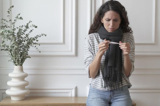 Unhealthy sick young Italian woman holding thermometer and measuring high temperature while standing indoors at home, unhappy worried female feeling unwell, suffering from flu, cold or covid-19