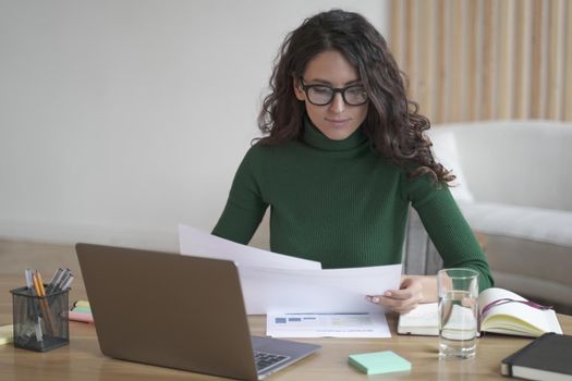Young focused Spanish woman freelancer in glasses analyzing documents, working on laptop computer at home, thoughtful Italian female sitting at desk, holding papers while preparing report