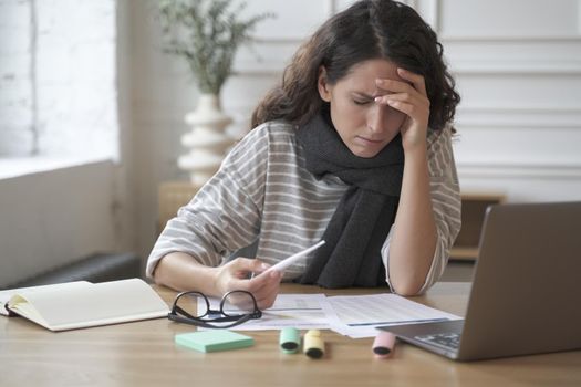 Young latin woman office worker sitting at table with digital thermometer in hand, suffering from headache and high fever at work, female emloyee feeling unwell sick, measuring temerature at workplace