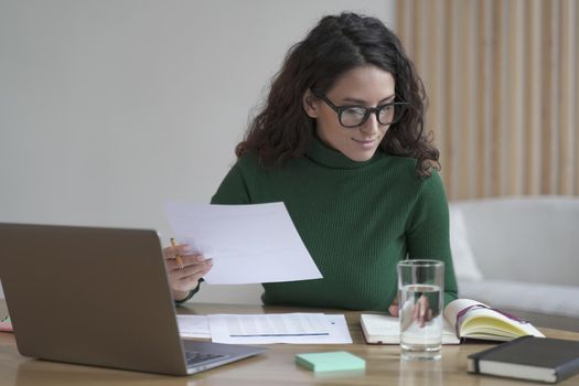Young pleasant Italian female wearing glasses holding documents preparing report while working on laptop computer in modern office, attractive Spanish woman in spectacles analyzing financial data