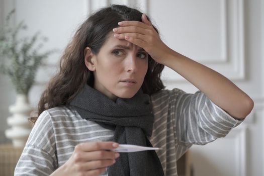 Sad sick young Italian woman wearing knitted scarf around neck suffering from flu cold while sitting at home with digital thermometer in hand, worried female touching head checking body temperature