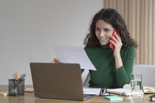 Pleasant young Italian businesswoman talking on mobile phone and consulting client, working remotely at home office, lookin at laptop screen with paper document in hand and making call on smartphone