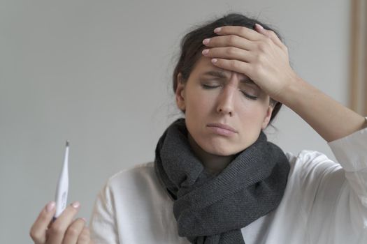 Italian entrepreneur lady tormented by hard work and fever, stands with closed eyes at home interior with warm scarf around her neck, while one hand on forehead and another holding digital thermometer