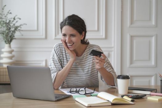 Happy exited Italian entrepreneur lady sitting at desk at home office while joyfully looking at computer screen holding credit card in hand, positive businesswoman making payments or shopping online