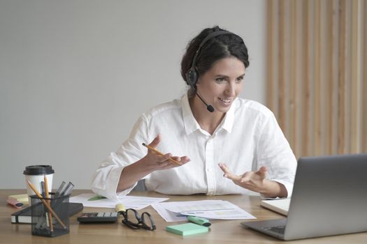 Smiling european woman online tutor sitting at desk in headset and talking by video call on laptop computer while working remotely in home office. Distance education and e-learning concept