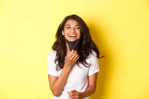 Concept of covid-19, social distancing and lifestyle. Image of beautiful african-american girl, feeling happy to breath freely after taking-off face mask, smiling pleased, yellow background.