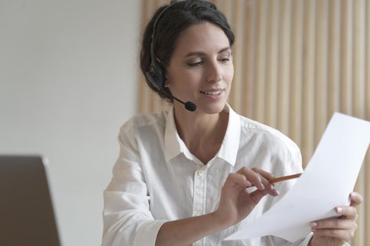 Portrait of positive focused Spanish business woman in headset sits at desk having video conference online with partner, holds accounting report in hand points to calculations in pencil