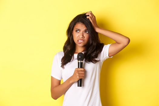 Image of indecisive african-american girl, holding microphone and looking hesitant, standing over yellow background.